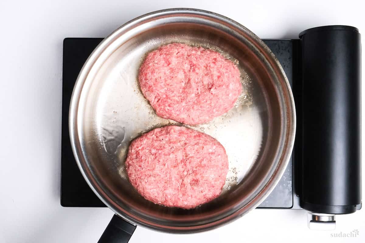 two hamburg patties frying in a stainless steel pan on the stove