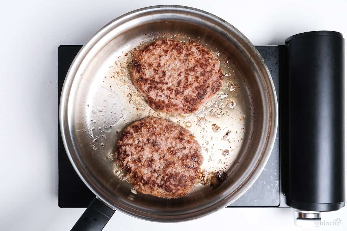 two hamburg patties frying in a stainless steel pan
