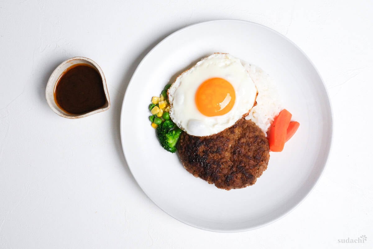 Japanese hamburg steak on a white plate with vegetables, rice and a fried egg with hamburg sauce in a small jug on the side