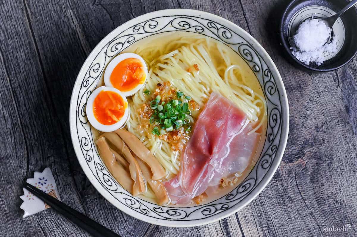 Homemade shio ramen topped with soft boiled eggs, menma, and ham in a cream bowl with black patterned rim on ashy wood background top down