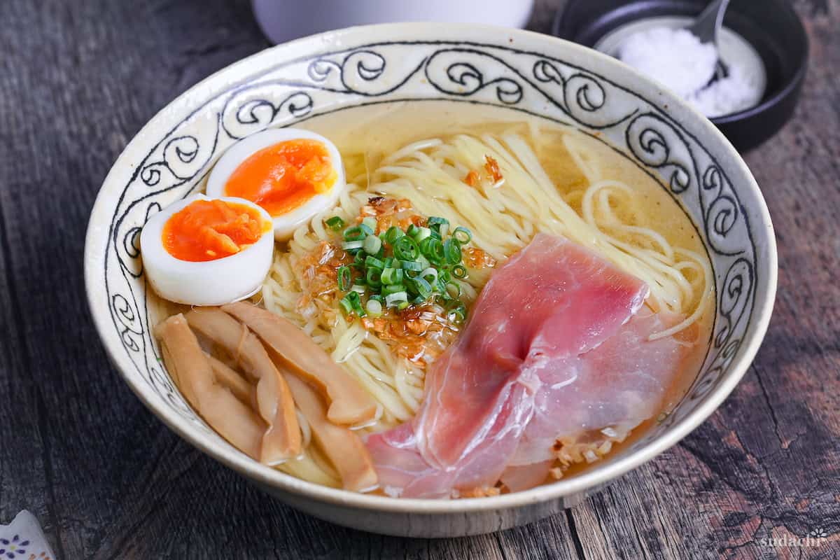 Homemade shio ramen topped with soft boiled eggs, menma, and ham in a cream bowl with black patterned rim on ashy wood background side view