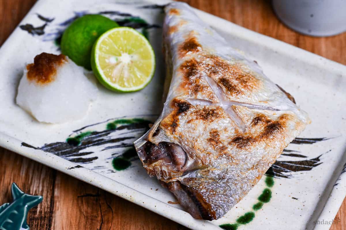 Salt Grilled Yellowtail Collar (Buri Kama Shioyaki) on a rectangular plate with halved sudachi and grated daikon drizzled with soy sauce