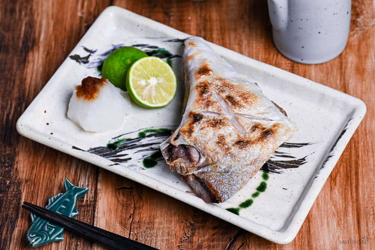 Salt Grilled Yellowtail Collar (Buri Kama Shioyaki) on a rectangular plate with halved sudachi and grated daikon drizzled with soy sauce