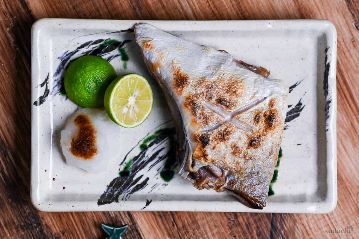 Salt Grilled Yellowtail Collar (Buri Kama Shioyaki) on a rectangular plate with halved sudachi and grated daikon drizzled with soy sauce