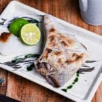 Salt Grilled Yellowtail Collar (Buri Kama Shioyaki) on a rectangular plate with halved sudachi and grated daikon drizzled with soy sauce