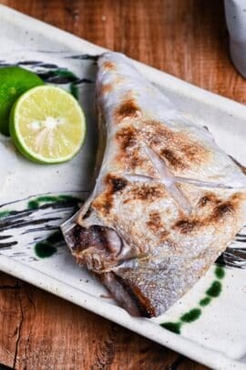 Salt Grilled Yellowtail Collar (Buri Kama Shioyaki) on a rectangular plate with halved sudachi and grated daikon drizzled with soy sauce