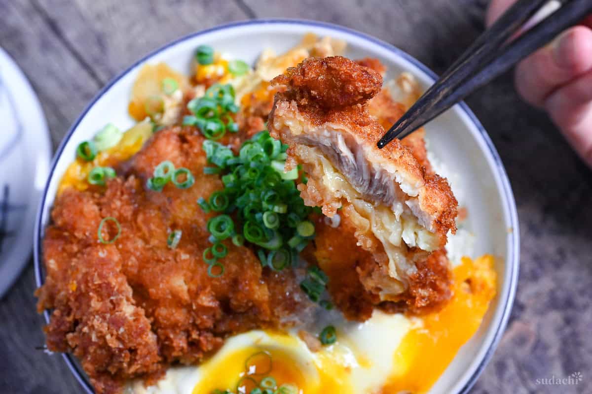 Black chopsticks holding up a piece of chicken cutlet over a bowl of chicken katsudon