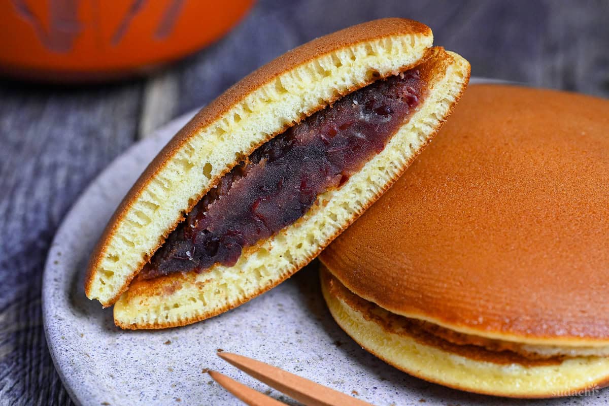 Half a dorayaki pancake revealing tsubuan (chunky red bean paste) in the center resting on a whole dorayaki on a mottled plate