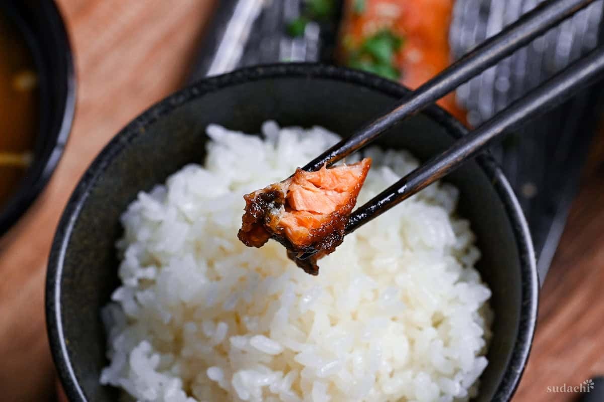 holding a piece of miso salmon with black chopsticks over rice
