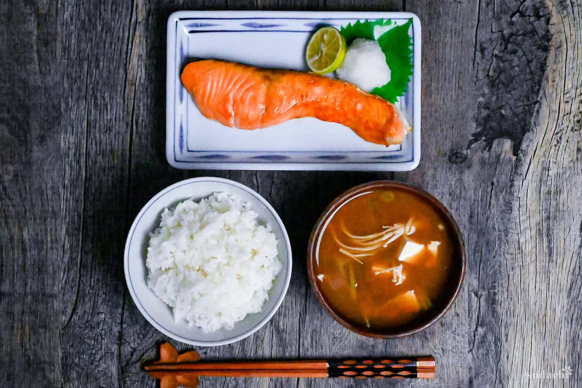 Japanese salt grilled breakfast salmon (shiozake) next to grated daikon and a sudachi on a white plate with blue rim with rice and miso soup