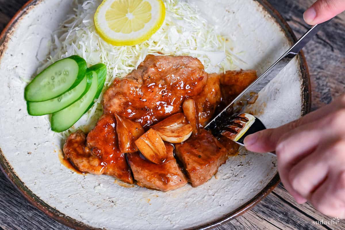 A knife and fork cutting into tonteki pork steak topped with garlic and a glossy sauce served with shredded cabbage, cucumber slices and a piece of lemon on a textured off-white plate with brown rim