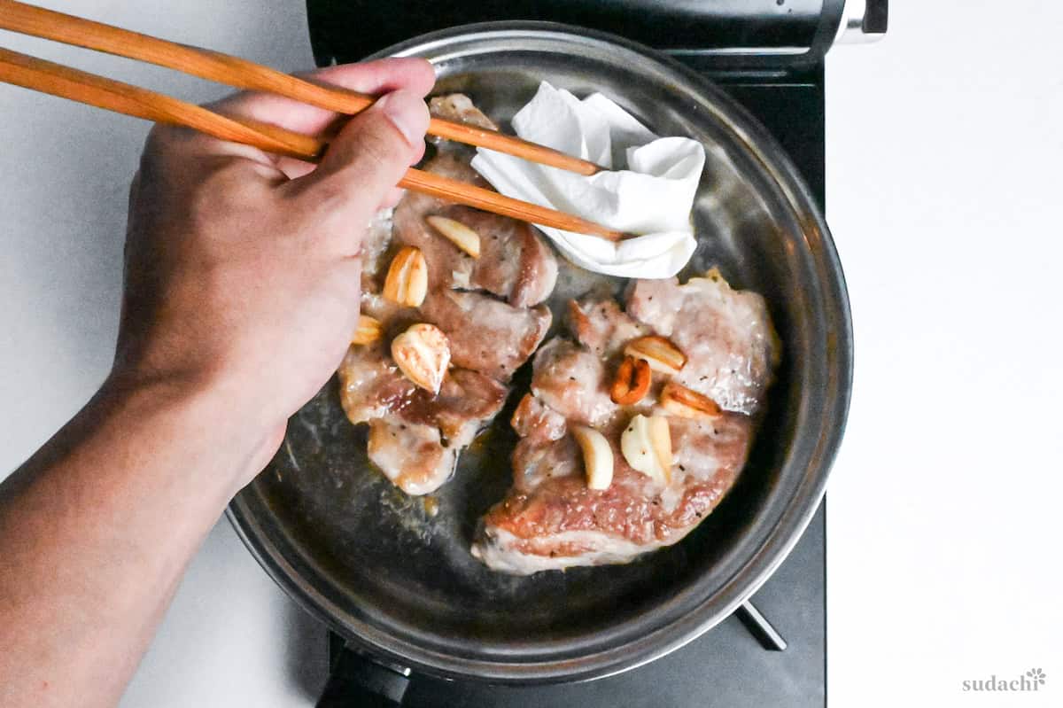 Using kitchen paper to wipe excess oil out of stainless steel pan with seared pork steaks and garlic