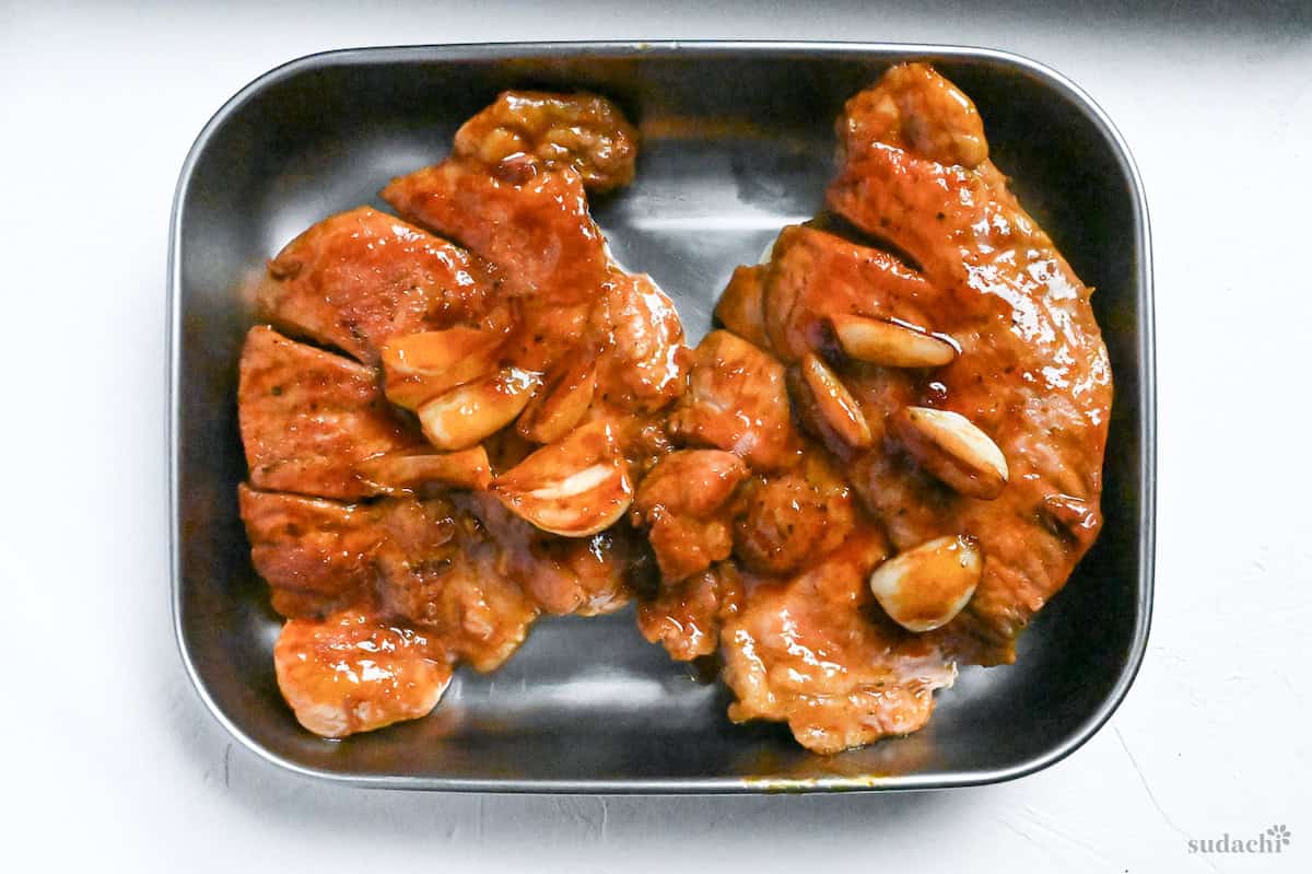 Two Japanese tonteki pork steaks topped with softened garlic in a steel container on a white surface