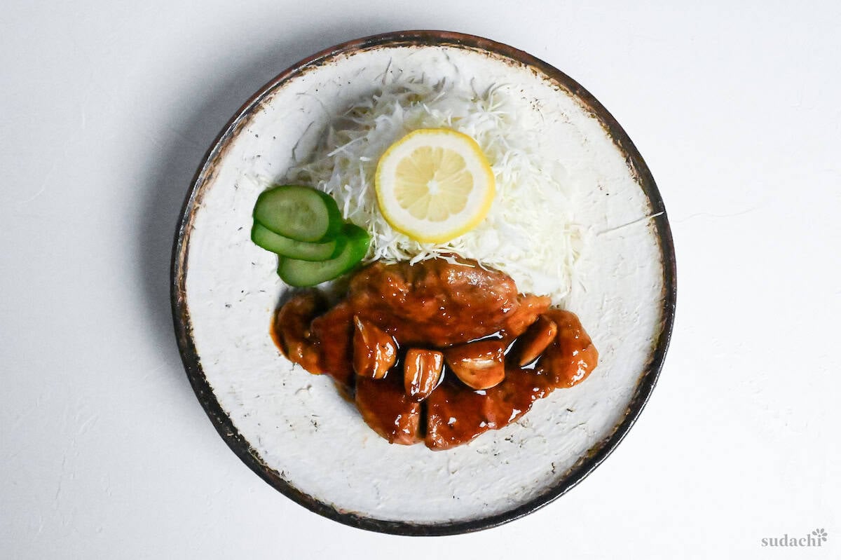 Tonteki pork steak with shredded cabbage, slices cucumbers and a piece of lemon on a white textured plate with brown rim