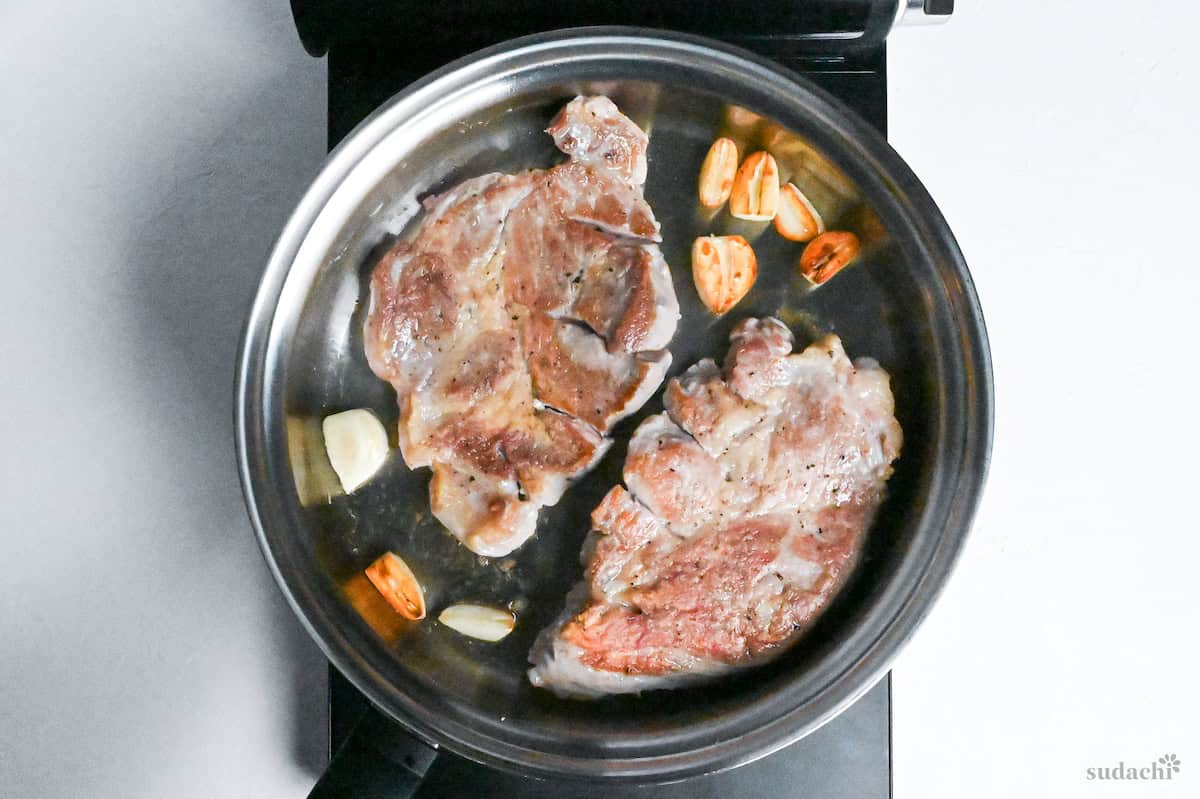 Browned pork steaks and garlic in a stainless steel pan on the stove