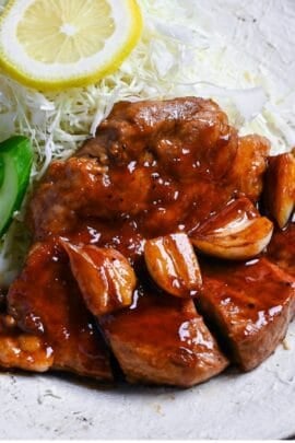 Close up of tonteki pork steak topped with garlic and a glossy sauce served with shredded cabbage, cucumber slices and a piece of lemon on a textured off-white plate with brown rim