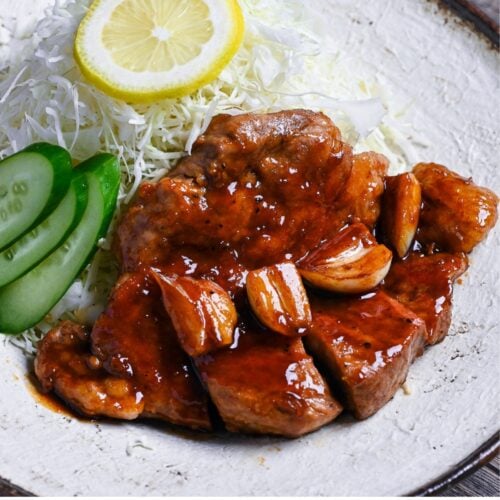 Close up of tonteki pork steak topped with garlic and a glossy sauce served with shredded cabbage, cucumber slices and a piece of lemon on a textured off-white plate with brown rim