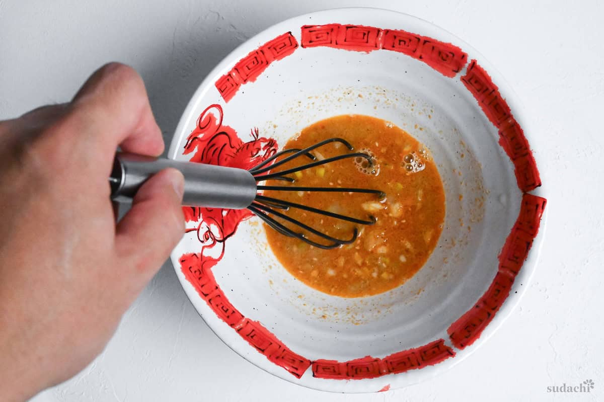 whisking tantanmen tare in a white ramen bowl with red dragon design rim