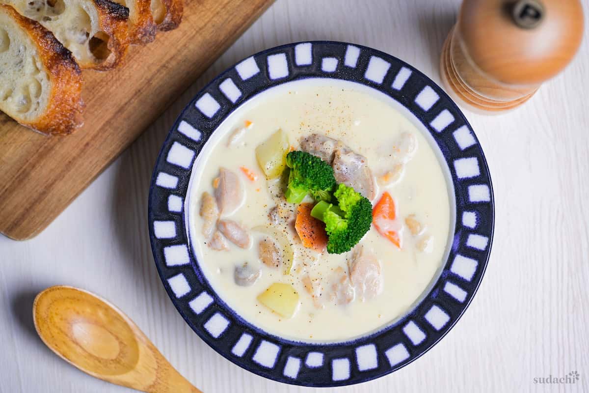 Japanese chicken cream stew in a white bowl with blue and white block patterned rim, next to sliced baguette and a light wooden spoon