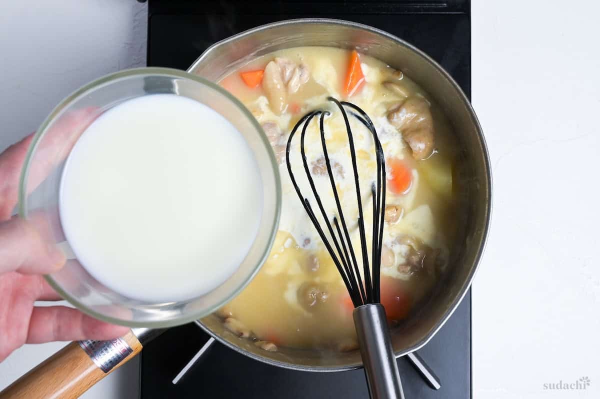 pouring cream and milk into chicken cream stew in a Japanese yukihira pot on the stove top with black silicone whisk