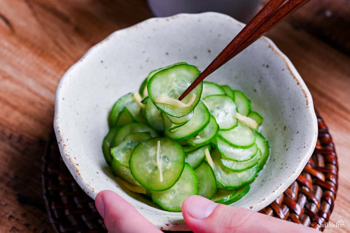 Japanese cucumber sunomono with ginger in a white mottled bowl with wooden chopsticks holding up a few slices