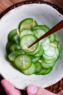 Japanese cucumber sunomono with ginger in a white mottled bowl with wooden chopsticks holding up a few slices