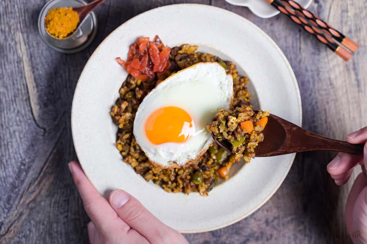 A spoonful of Japanese style curry fried rice held above a white bowl of curry fried rice topped with a fried egg and some Japanese curry pickles (fukujinzuke)