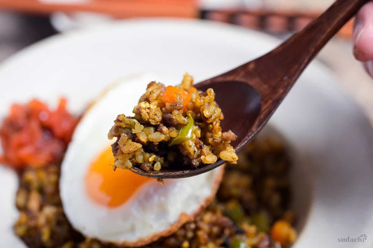 A spoonful of Japanese style curry fried rice held above a white bowl of curry fried rice topped with a fried egg and some Japanese curry pickles (fukujinzuke)