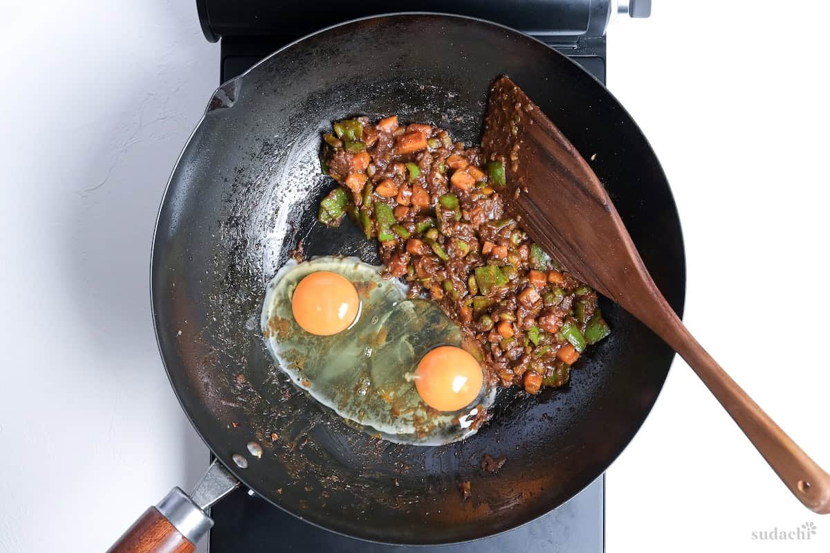 Cury rice ingredients pushed to one side in a wok, with two eggs cracked into the empty space