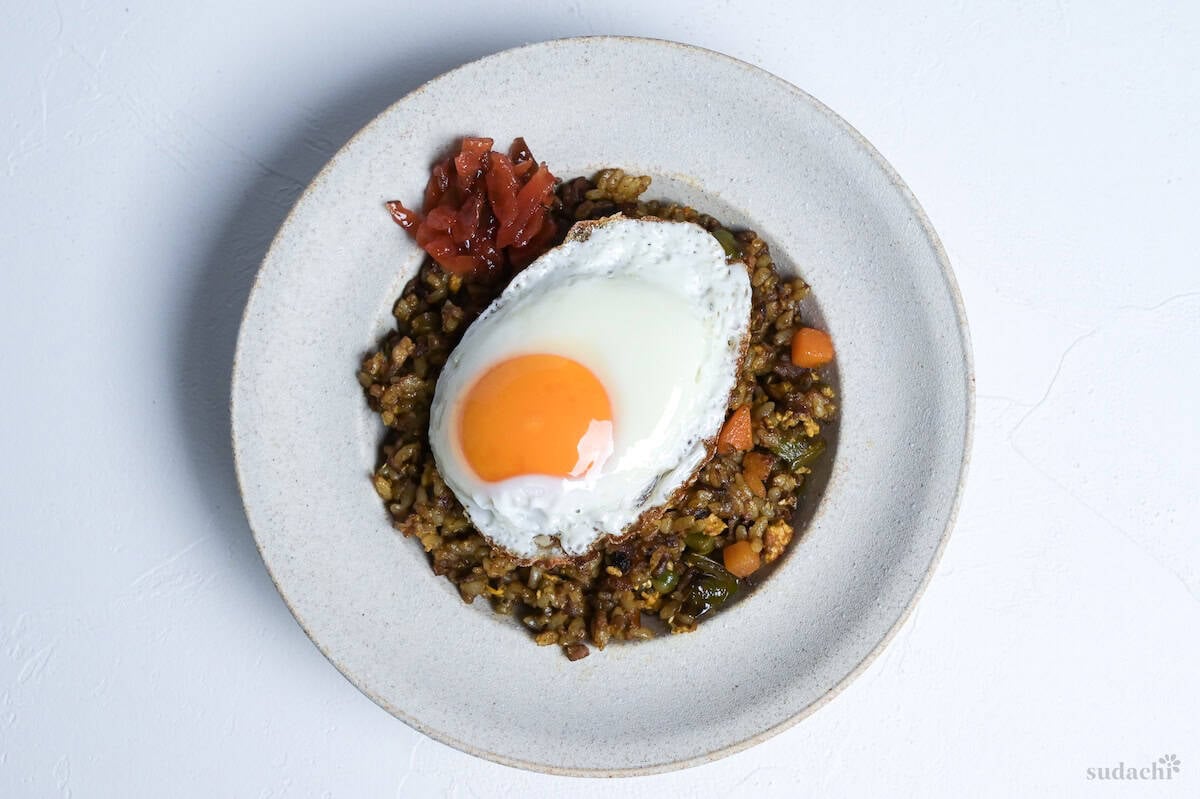 Japanese style curry rice on an off-white plate topped with a sunny-side up egg and some fukujinzuke pickles