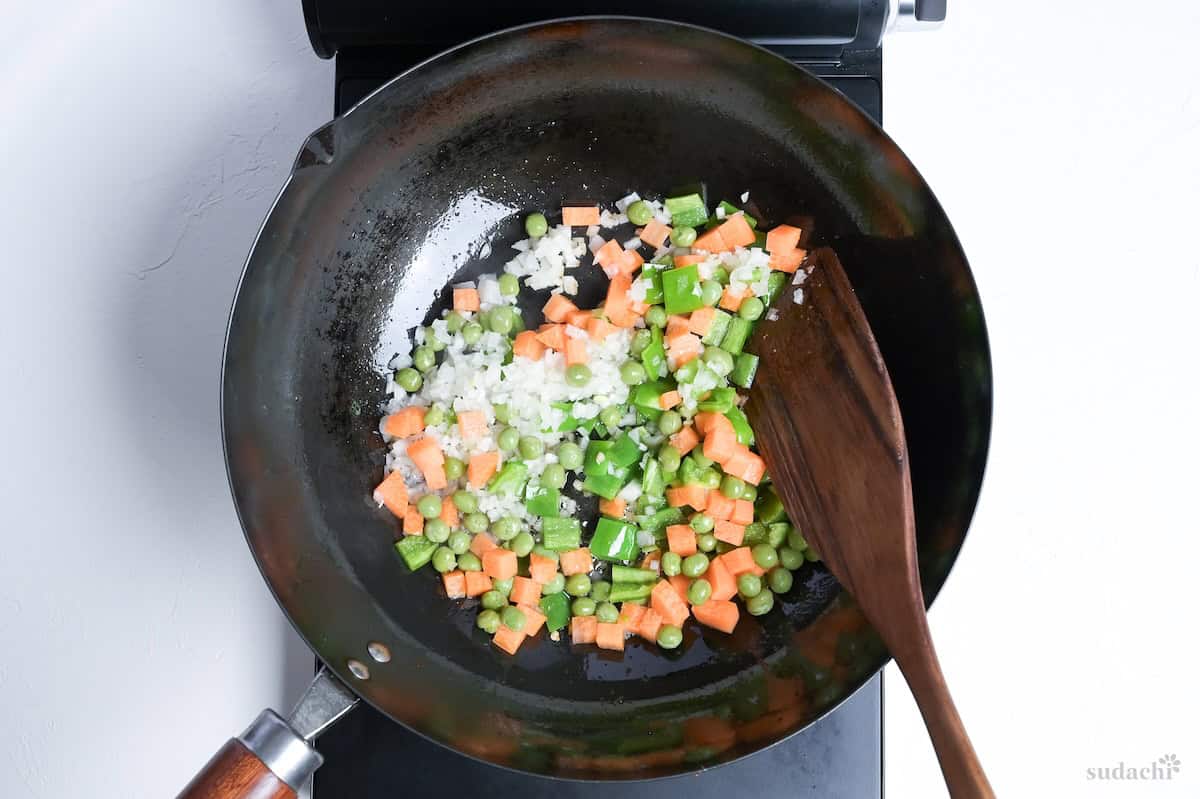 finely diced onion, carrot, green bell peppers and green peas in a wok with a dark wooden spatula