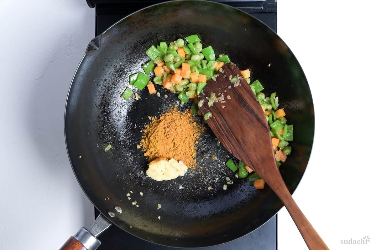 Vegetables in a wok pushed to one side and grated garlic & ginger and curry powder in the empty space in the pan
