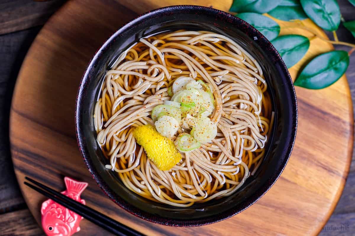 Kake Soba Noodle Soup in a black and red Japanese bowl topped with chopped green onions, yuzu peel and a sprinkle of shichimi togarashi (Japanese 7 spice chili powder) top down