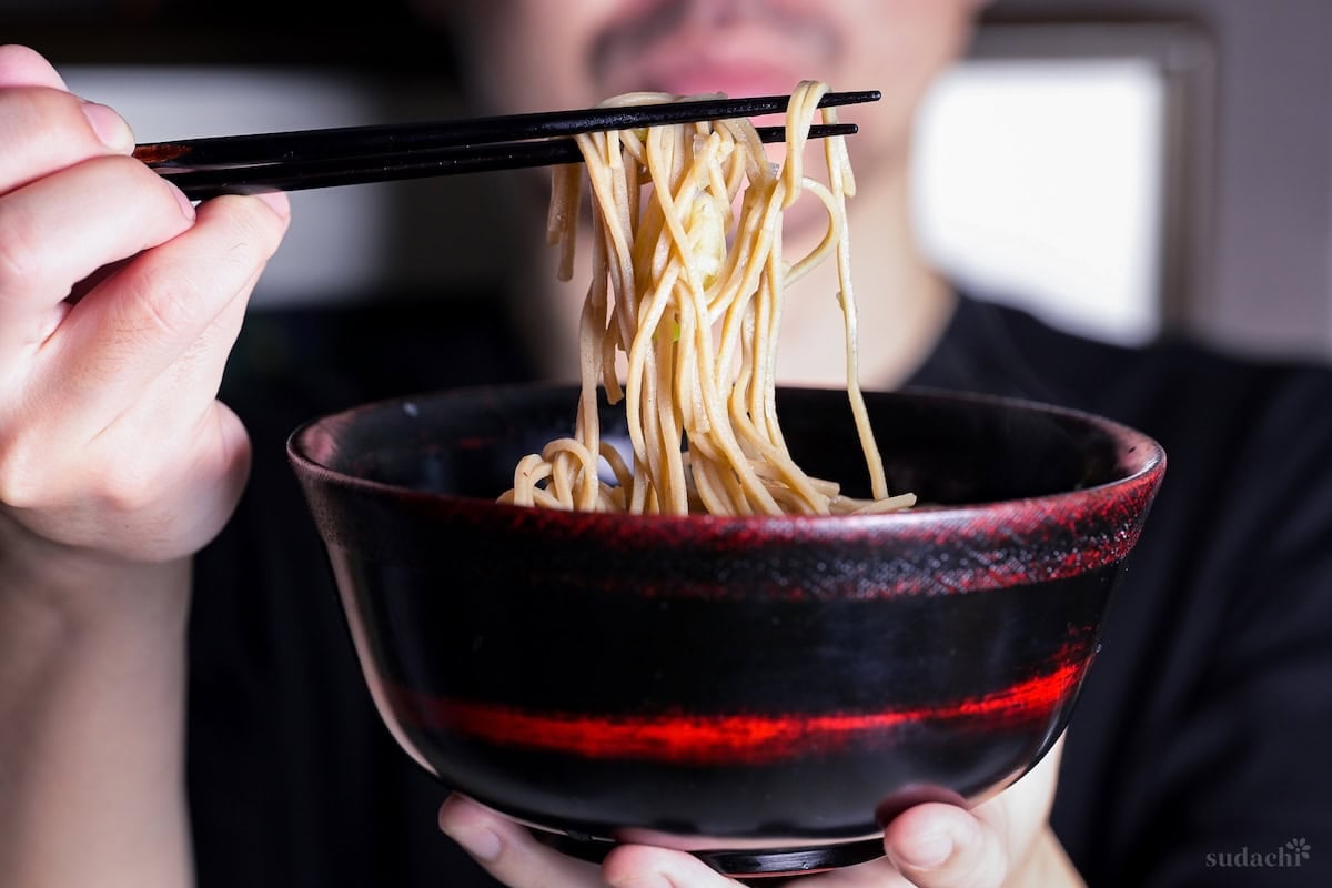 Yuto Omura holding up a black and red bowl of kake soba with black chopsticks holding up soba noodles