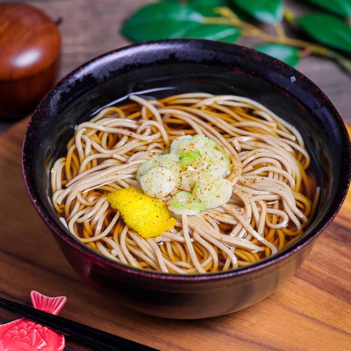 Kake Soba Noodle Soup in a black and red Japanese bowl topped with chopped green onions, yuzu peel and a sprinkle of shichimi togarashi (Japanese 7 spice chili powder) side view
