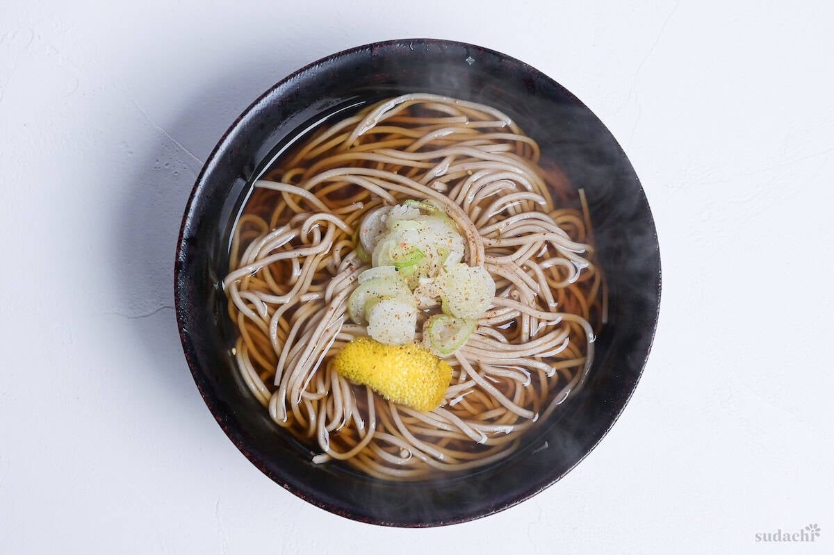 Kake Soba noodle soup in a black bowl topped with topped green onions, yuzu peel and a sprinkle of shichimi togarashi (Japanese 7 spice chili powder)