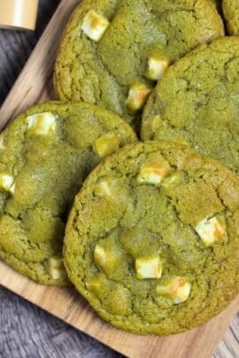 6 matcha and white chocolate cookies on a wooden cutting board next to matcha powder in a small brown dish and a bamboo matcha whisk (chasen)
