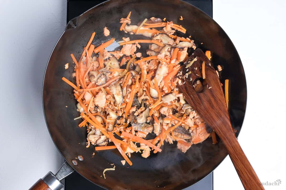Salmon fillets broken into flakes and mixed with thinly sliced carrots, mushrooms and ginger in a wok on the stove