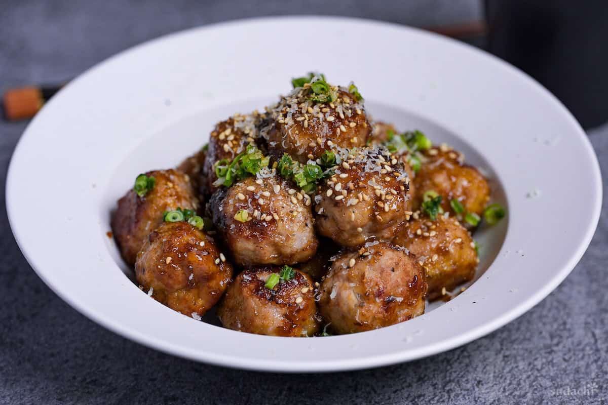 Teriyaki meatballs stacked in a pyramid on a white dish and topped with sesame seeds, chopped green onions and grated parmesan cheese