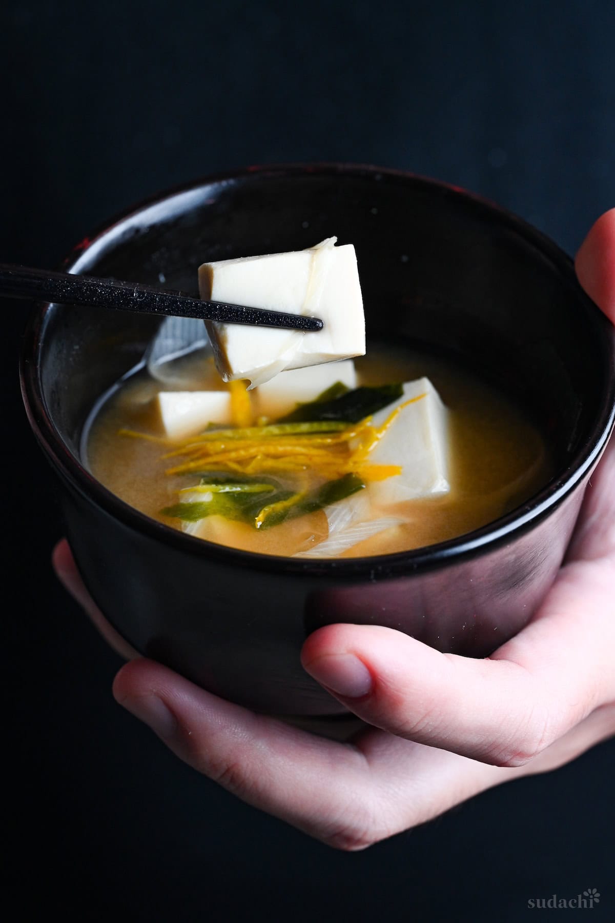 Yuto Omura holding Japanese tofu miso soup in a black bowl in one hand, and holding a piece of tofu with black chopsticks with the other