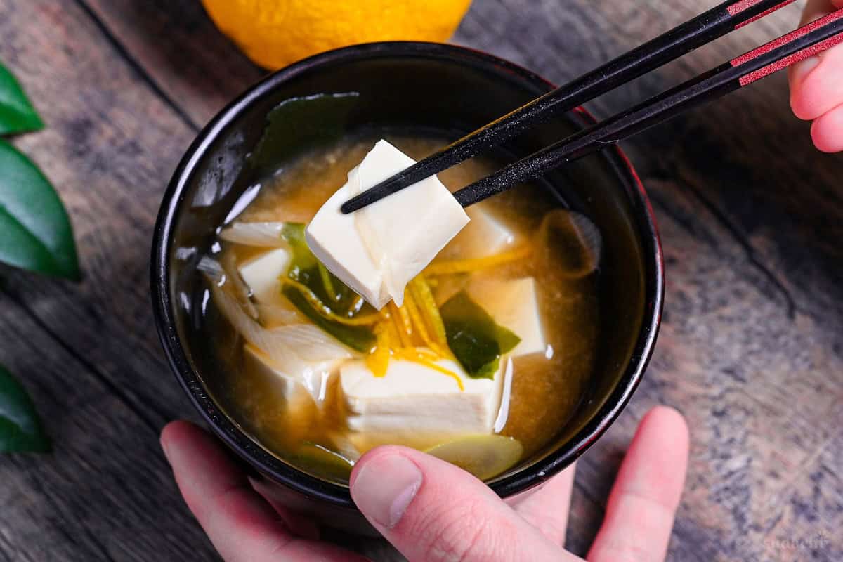 Japanese tofu miso soup in a black bowl with black chopsticks holding a piece of silken tofu above the bowl