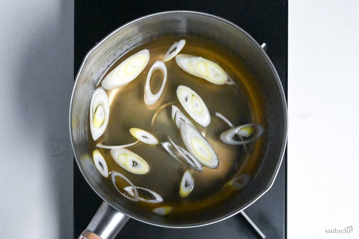 Diagonally sliced pieces of Japanese leek simmering in dashi in a pot on the stove top