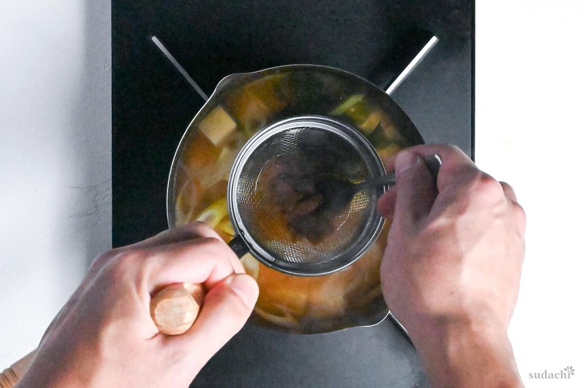 Adding miso to dashi broth using a miso strainer and metal spoon