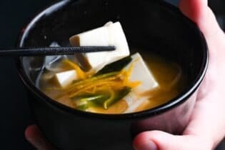 Yuto Omura holding Japanese tofu miso soup in a black bowl in one hand, and holding a piece of tofu with black chopsticks with the other, thumbnail size
