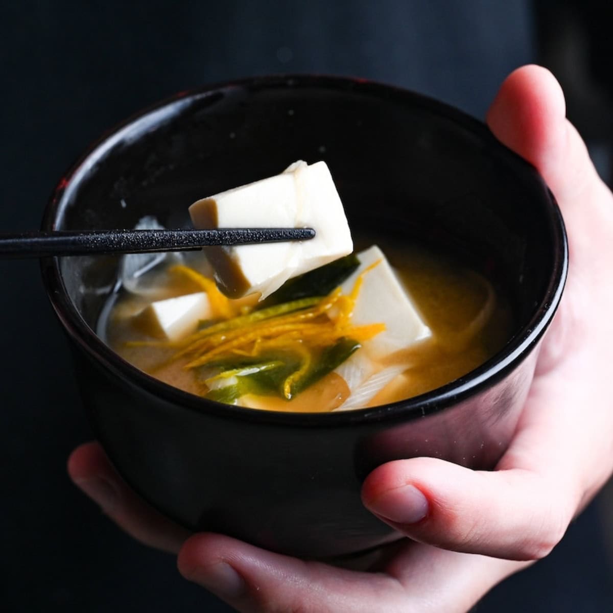 Yuto Omura holding Japanese tofu miso soup in a black bowl in one hand, and holding a piece of tofu with black chopsticks with the other, thumbnail size