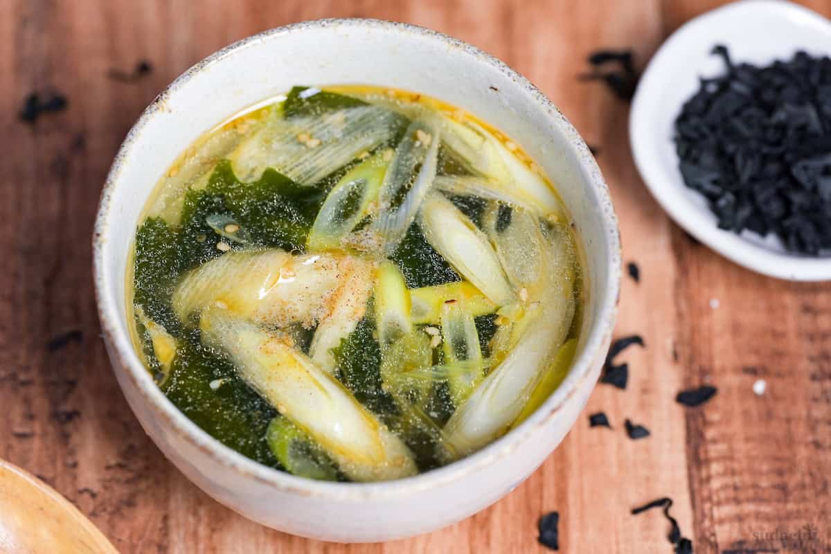 Wakame soup in an off-white ceramic bowl next to a small bowl of dried wakame and a light wooden spoon