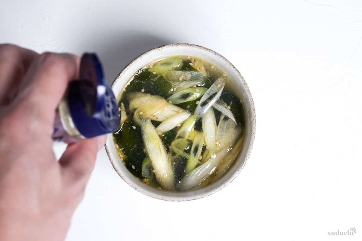 Wakame soup with Japanese leek in an off-white ceramic bowl, with a hand holding ground white pepper above it