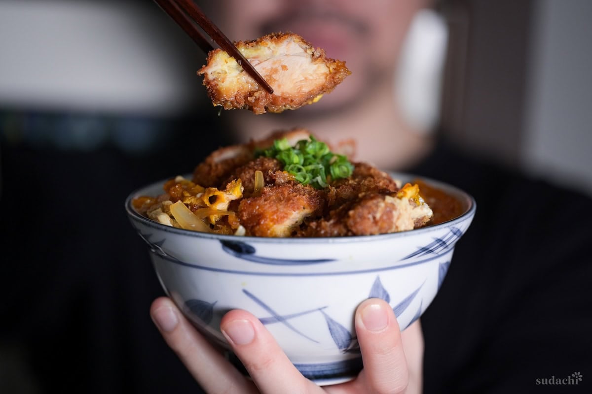Yuto Omura holding up a bowl of chicken katsudon in one hand, with chopsticks holding a piece of chicken katsu in the other