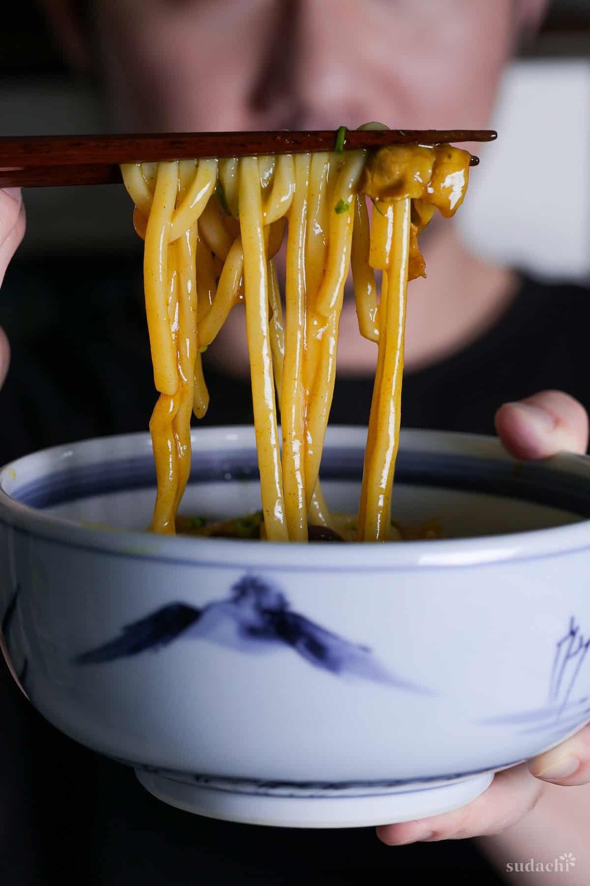 Yuto Omura holding a white and blue bowl of curry udon in one hand, with dark wooden chopsticks holding up udon in the other hand
