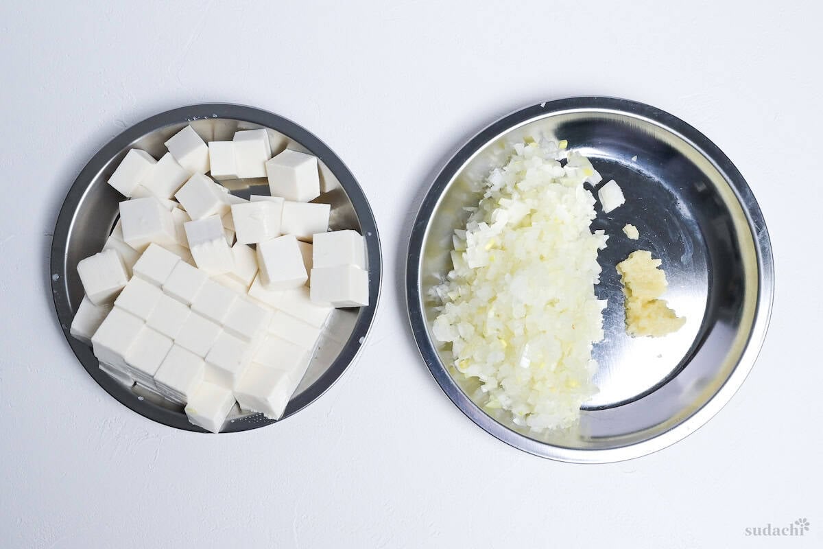 Cubed firm tofu, finely diced onion and grated garlic on steel prep plates on a white background